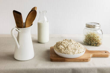 Kanne mit Holzlöffel und Spachtel, Milchflasche, Glas mit Sprossen und Teig auf dem Schneidebrett - EVGF03707