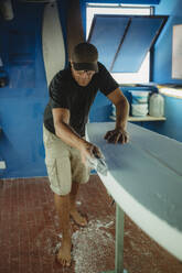 Qualifizierter Arbeiter glättet barfuß ein weißes Surfbrett in einer Werkstatt mit blauen Wänden - ADSF13133
