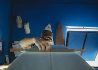 Konzentrierter Facharbeiter mit Schutzmaske beim Ausrichten eines weißen Surfbretts in einer Werkstatt mit blauen Wänden - ADSF13135