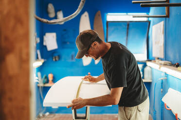 Seitenansicht eines erwachsenen Mannes, der ein weißes Brett ausmisst, während er in einer kleinen Werkstatt mit blauen Wänden arbeitet und ein Surfbrett herstellt - ADSF13136