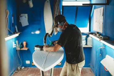 Arbeiter mit Schutzmaske beim Einstellen von Details am Surfbrett in der Werkstatt - ADSF13142