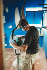 Arbeiter mit Schutzmaske beim Einstellen von Details am Surfbrett in der Werkstatt - ADSF13143