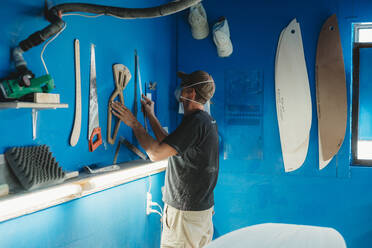 Seitenansicht eines Handwerkers mit Schutzmaske, der ein Instrument von der blauen Wand nimmt, während er in der Werkstatt arbeitet und Surfbretter herstellt - ADSF13144