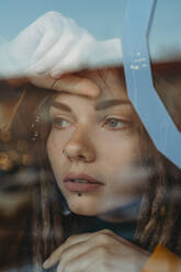 Nachdenkliche, traurige junge Frau mit Dreadlocks, die sich an eine Fensterscheibe lehnt und wegschaut - ADSF13152