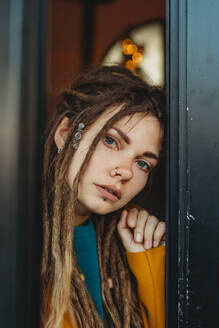 Stilvolle Millennial-Frau mit Dreadlocks, die einen gelben Mantel über einem blauen Pullover trägt und nachdenklich wegschaut, während sie in einem Raum mit einem Klavier im Hintergrund sitzt - ADSF13153