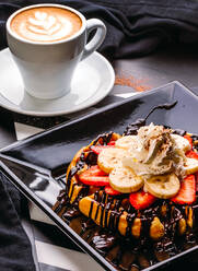 Von oben Cappuccino in weißem Becher auf Tisch mit Teller mit runder Waffel mit Banane und Erdbeere, gekrönt mit Schokoladensauce und Schlagsahne - ADSF13179