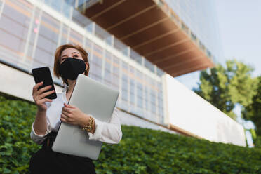 Niedriger Winkel einer entschlossenen weiblichen Führungskraft mit medizinischer Maske, die mit Laptop und Smartphone auf der Straße steht und wegschaut - ADSF13192