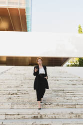 Content female manager in elegant suit walking down the stairs in city and on cellphone while looking away - ADSF13195