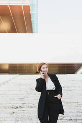 Content female manager in elegant suit walking down the stairs in city and discussing work issues on cellphone while looking away - ADSF13196