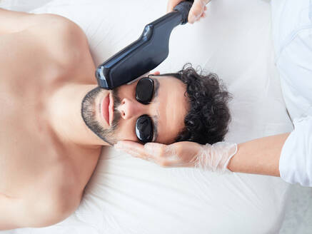 Von oben abgeschnittenen gesichtslosen Meister mit Laser-Gerät auf männlichen Gesicht entfernen Bart Gesicht während des Verfahrens in Salon - ADSF13235