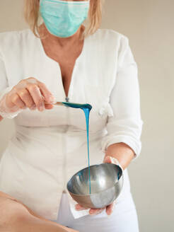Gesichtslose Frau in medizinischer Maske und Handschuhen rührt heißes blaues Wachs mit einem Holzstab in einer Schüssel - ADSF13236