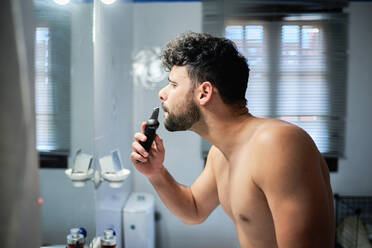 Side view of serious shirtless handsome male using electric shaver for grooming while standing in modern bathroom and looking in mirror - ADSF13240