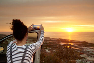 Rückansicht einer nicht erkennbaren weiblichen Touristin, die in der Nähe eines Autos steht und mit ihrem Handy ein Foto von einem wunderschönen Sonnenuntergang über dem Meer macht - ADSF13244