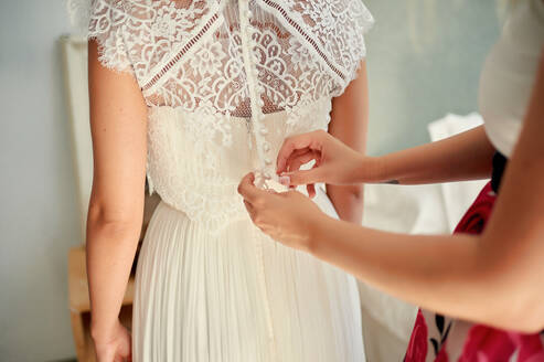 Unerkannte Brautjungfer hilft eleganter Braut beim Zuknöpfen des weißen Kleides, während sie in einem hellen Raum zu Hause steht - ADSF13245