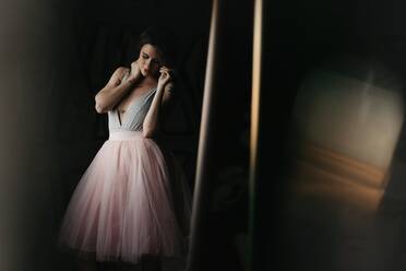 Calm graceful young female dancer in elegant gown touching hair gently while standing looking away in dark studio - ADSF13250