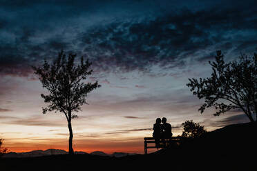 Rückenansicht Silhouetten von liebevollen Paar genießen romantische Datum beim Sitzen auf der Bank gegen bewölkten Sonnenuntergang Himmel im Sommer Abend - ADSF13255