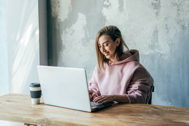 Junge weibliche Remote-Mitarbeiterin, die an einem Netbook arbeitet, während sie an einem Holztisch mit einem Einwegglas Kaffee in der Nähe einer alten Zementwand in einer Wohnung sitzt - ADSF13265