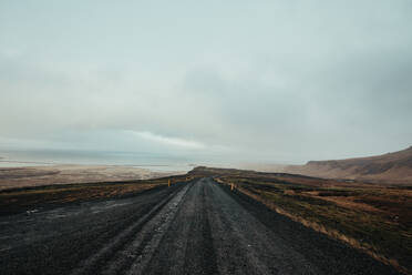 Majestätischer Blick auf eine schmale, leere, gerade Asphaltstraße, umgeben von Bergen unter einem Himmel mit niedrigen Wolken bei Tageslicht in Island - ADSF13267