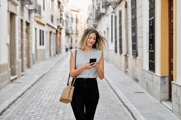 Glückliche junge Frau in stilvollem Outfit, die mit ihrem Handy telefoniert, während sie auf einer alten, engen Straße in der Stadt spazieren geht und wegschaut - ADSF13273