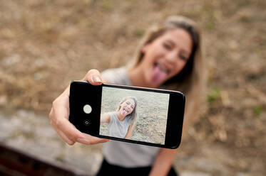 Hoher Winkel der spielerischen jungen Frau macht Gesicht und zeigt Zunge bei der Aufnahme von Selfie auf Smartphone im Sommer Tag auf der Straße - ADSF13275