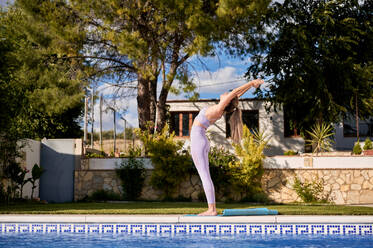 Seitenansicht einer schlanken Frau in Sport-BH und Leggings, die auf einer Matte am Pool steht und Yoga in Urdhva Hastasana praktiziert - ADSF13279