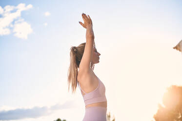 Niedriger Winkel Seitenansicht der heiteren weiblichen stehend in Urdhva Hastasana mit erhobenen Armen in Namaste Geste und üben Yoga während des Sonnenuntergangs im Park - ADSF13286