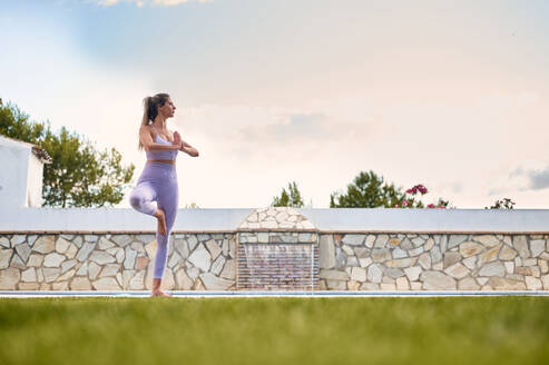 Ruhige Frau in Sportleggings und BH balanciert auf einem Bein in Vrksasana mit Namaste-Geste, während sie Yoga macht und wegschaut - ADSF13287