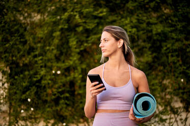 Schlanke, nachdenkliche Frau in Sportkleidung, die mit einer gerollten Yogamatte steht, während sie in der Nähe von Bäumen ein Mobiltelefon benutzt und bei Tageslicht wegschaut - ADSF13290