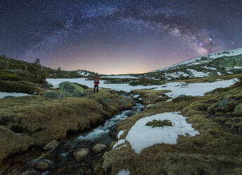 Back view of unrecognizable distant hiker standing near small river streaming through mountainous terrain covered with snow and enjoying spectacular scenery with Milky Way in dark sky in spring night - ADSF13296