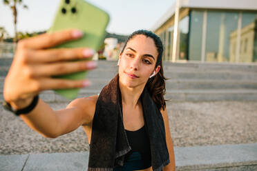 Junge schlanke Frau in Sportkleidung mit Ohrhörern und Fitness-Tracker, die ein Selfie mit ihrem Handy macht, während sie nach dem Training auf der Straße steht - ADSF13313