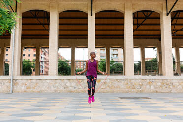 Full body of African American fit female athlete in colorful sportswear jumping rope on pavement in town near building while warming up - ADSF13315