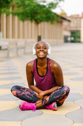 Positive afroamerikanische Sportlerin in Sportkleidung und Kopfhörern, die mit gekreuzten Beinen auf einer Böschung in der Nähe eines Gebäudes in der Stadt sitzt und in die Kamera schaut, während sie Musik hört - ADSF13319