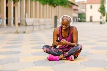 Positive afroamerikanische Sportlerin in Sportkleidung und Kopfhörern, die mit gekreuzten Beinen auf einer Böschung in der Nähe eines Gebäudes in der Stadt sitzt und auf ihrem Smartphone surft, während sie Musik hört - ADSF13320