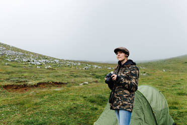 Positive junge Frau in Oberbekleidung mit professioneller Fotokamera, die in der Nähe eines Campingzelts in herbstlicher Landschaft steht und wegschaut - ADSF13323