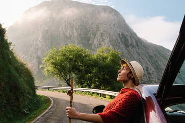 Seitenansicht einer sorglosen weiblichen Reisenden, die sich auf der Straße an ein Auto lehnt, vor dem Hintergrund einer spektakulären Landschaft im Hochland - ADSF13342