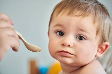 Nahaufnahme eines niedlichen kleinen Babys, das Püree von einem Löffel isst, der von einer anonymen Mutter gehalten wird - ADSF13359