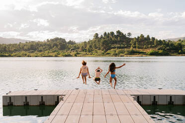 Rückenansicht von Kindern in Badekleidung beim Sprung in den Teich vom hölzernen Kai am Wochenende im Sommer - ADSF13379