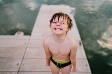 Hoher Winkel eines fröhlichen Kindes in Badekleidung und mit nassen Haaren, das auf einem Holzsteg steht und nach dem Schwimmen im Teich ein lustiges Gesicht macht - ADSF13382