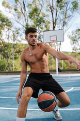 Concentrated male athlete with naked torso playing basketball alone on sports ground in summer - ADSF13383