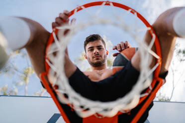 Männlicher Sportler in Sportkleidung, der auf einem Basketballkorb mit Ball sitzt und in die Kamera schaut, von unten - ADSF13384