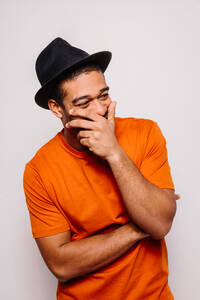 Fröhlicher Mann isoliert auf weißem Hintergrund, er trägt ein orangefarbenes T-Shirt und einen schwarzen Hut und schaut mit der Hand auf dem Mund weg - ADSF13401
