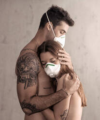 Seitenansicht eines nackten, tätowierten Freundes und einer Freundin, die sich umarmen, während sie während der Pandemie COVID 19 Schutzmasken tragen - ADSF13404