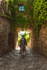 Rückenansicht einer nicht erkennbaren Frau in Kleid und Hut, die durch die Straßen eines mittelalterlichen Dorfes mit Kletterpflanzen in Europa geht - ADSF13426