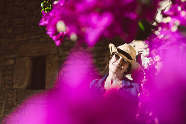 Woman wearing a hat looking at camera while standing near a wall with pink flowers shrubs - ADSF13428