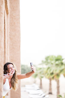 Fröhliche Frau, die ein Handy benutzt, um ein Selfie in der Nähe der Wüstenlandschaft auf einem steinernen Balkon zu machen, Marokko - ADSF13464