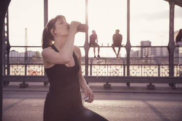 Frau trinkt Wasser aus einer Flasche, nachdem sie auf einer Brücke bei Sonnenuntergang in der Stadt trainiert hat - DHEF00316