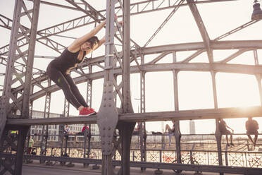 Woman stretching while exercising on metal bridge in city at sunset - DHEF00327