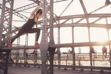 Determined woman stretching and exercising on railing of bridge at sunset - DHEF00328