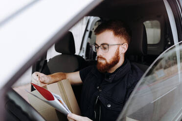Seitenansicht eines aufmerksamen, nachdenklichen erwachsenen Mannes, der sich auf Dokumente konzentriert und diese überprüft, während er hinter dem Lenkrad im Auto sitzt, auf einem unscharfen Hintergrund - ADSF13481