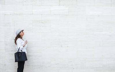 Side view of elegant female in stylish outfit and hat holding black bag with white wall on background - ADSF13496